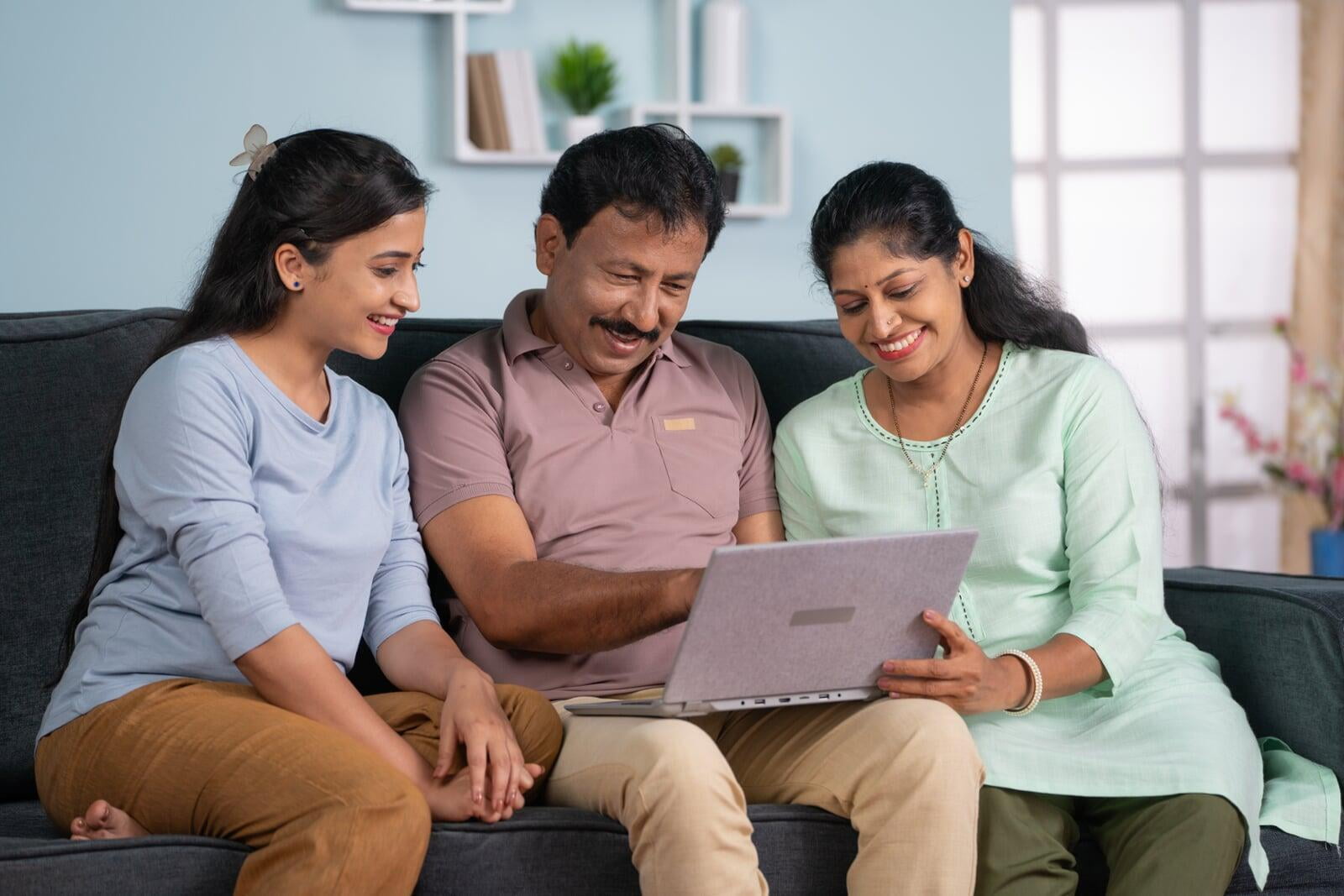 Happy indian middle aged parents with adult daughter using laptop on sofa at home